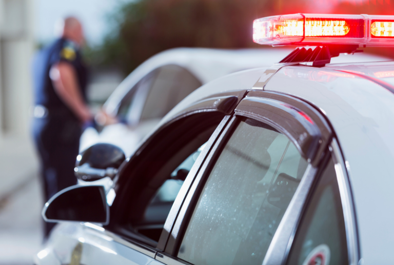 Image is of a police car with it's lights on, and in the background a police officer is next to a vehicle speaking to the driver, concept of felony DUI charges in Florida