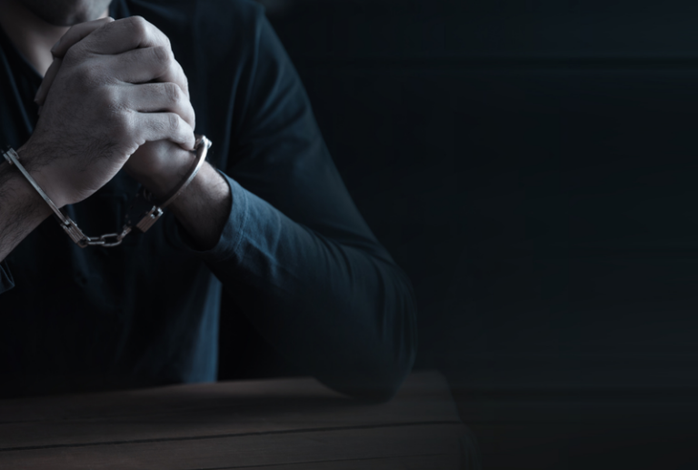 Image is of a man in a dark room sitting at a table with his hands cuffed on the table, concept of what is sexual assault?