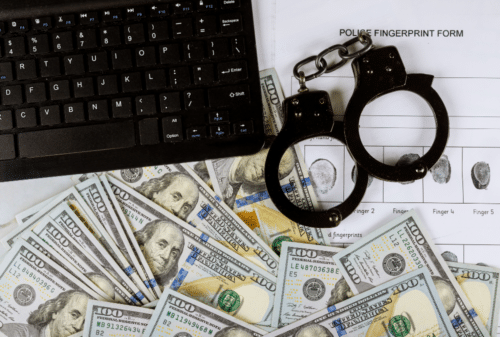 Image is of a keyboard, pile of hundred dollar bills, handcuffs, and finger print paper, concept of punishments for white-collar crime.