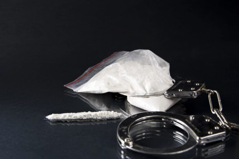 Image shows a bag of white powder and handcuffs placed on a dark surface, symbolizing arrest and charges under drug trafficking laws in Florida.