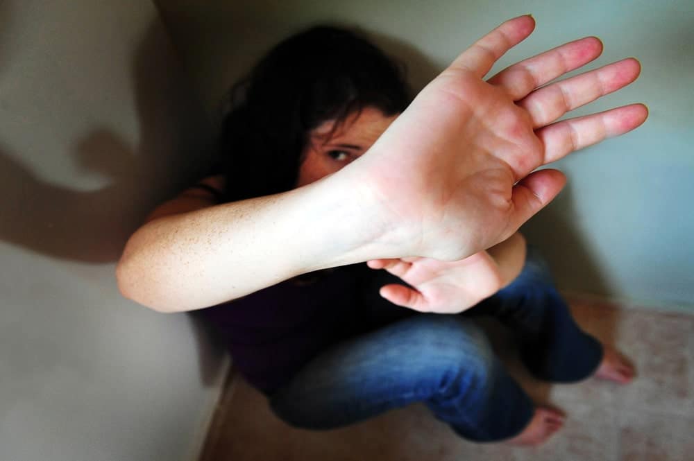 Image shows a woman sitting on the floor and shielding her face with her hand, illustrating the human impact behind domestic violence charges.
