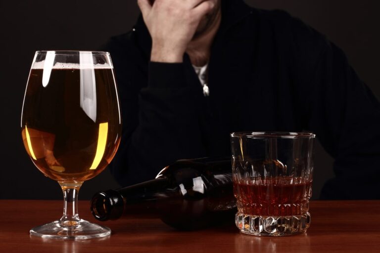 Image is of beer and liquor glasses on a table with a person in the background, concept of DUI and impaired decision making affecting professional licenses.
