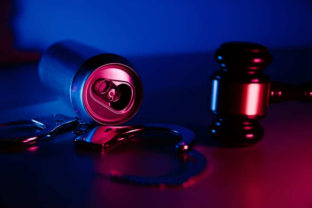 Image is of handcuffs, an alcoholic beverage can, and a judge's gavel under emergency lighting, concept of prior DUIs resulting in stricter penalties and stronger prosecution efforts.
