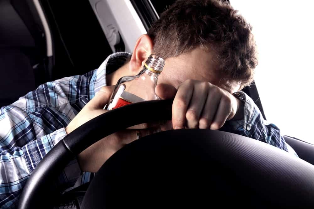 Image is of a driver holding an alcohol bottle behind the steering wheel, concept of conduct that can lead to DUI charges.