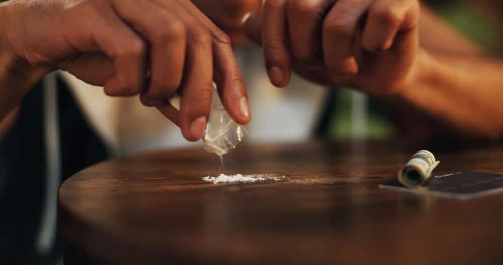 Image is of a person pouring white powder from a small plastic bag onto a table, representing evidence involved in drug charges without possession in Ponte Vedra Beach.